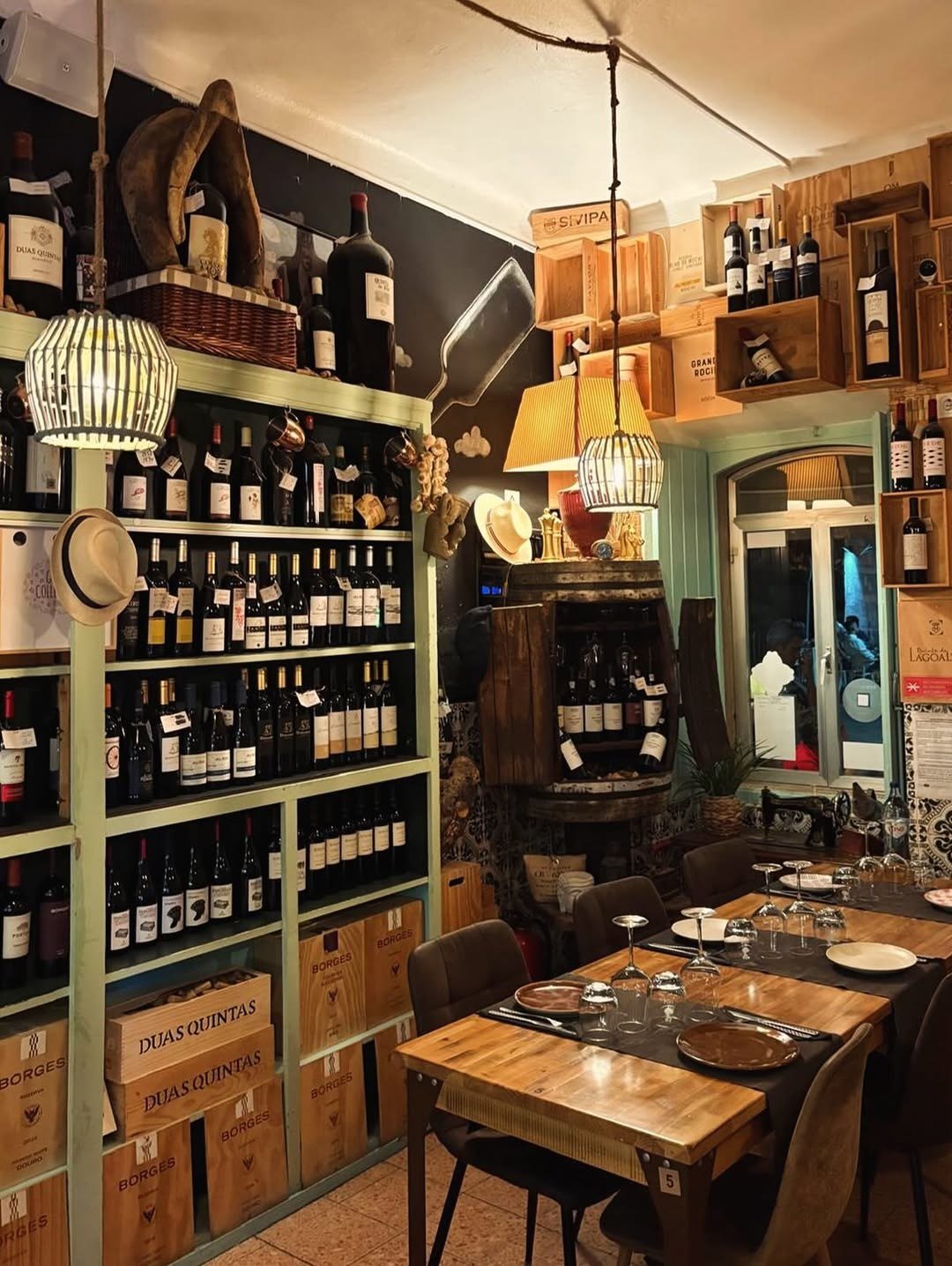 Sala com estantes de madeira cheias de garrafas de vinho, candeeiro de vime e mesas postas