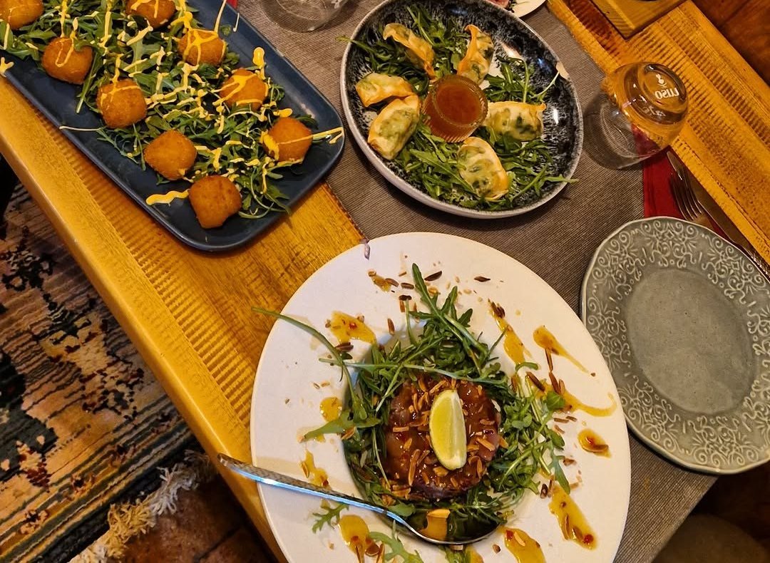 Mesa de madeira vista de cima com vários pratos: bolinhas fritas sobre rúcula, pastéis com molho e um prato com rúcula e lima