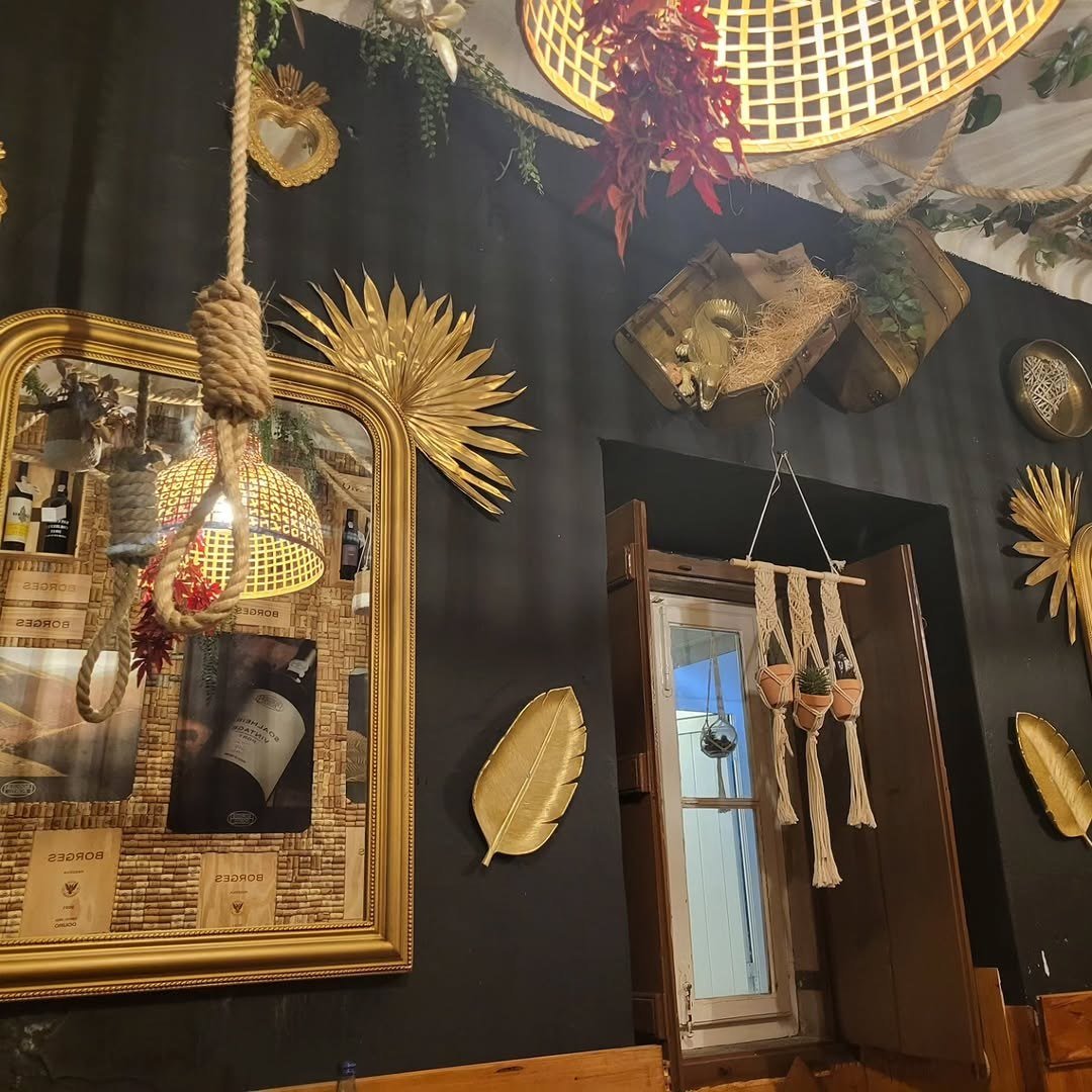 Sala de paredes escuras com espelho dourado, candeeiro de vime, plantas e garrafas de vinho